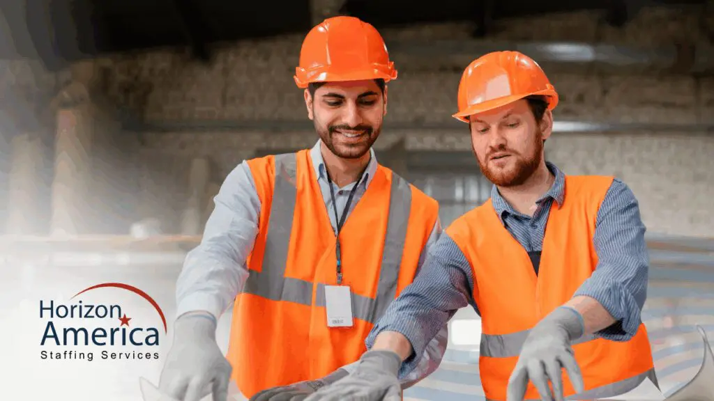 Two light industrial workers wearing safety vests and hard hats collaborate onsite as part of light industrial workforce management.