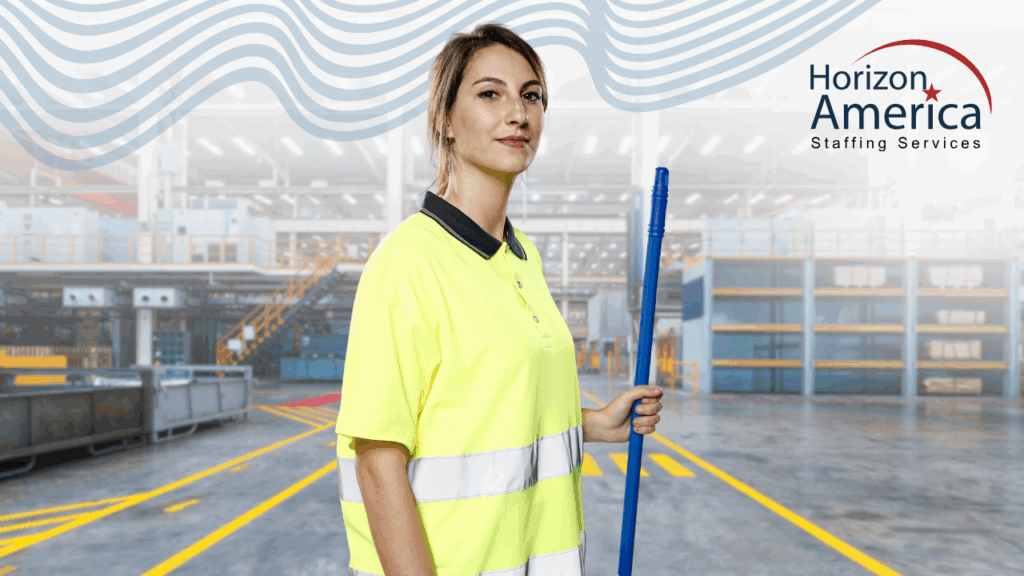 Worker in sanitation services stands holding a long-handled cleaning tool on an industrial work floor.