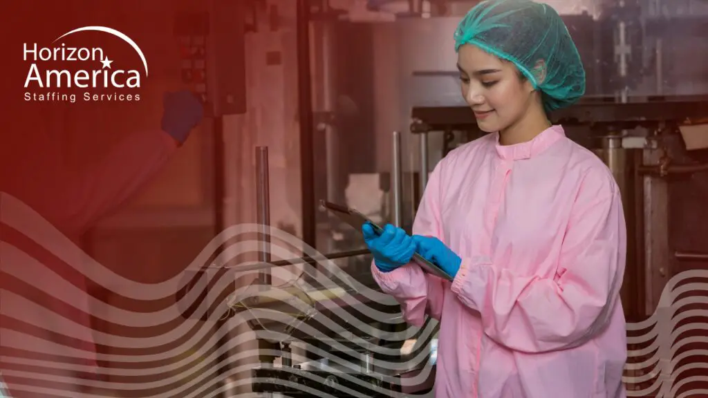 A food processing worker in proper protective gear, hairnet, and gloves works on a tablet.