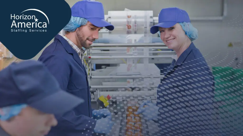 Two food manufacturing workers in blue uniforms, caps, and gloves work side by side on a production line inside a food processing facility.
