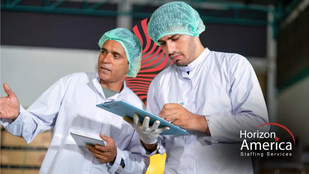 Two food manufacturing supervisors in white lab coats and hairnets review a clipboard together on a production floor.