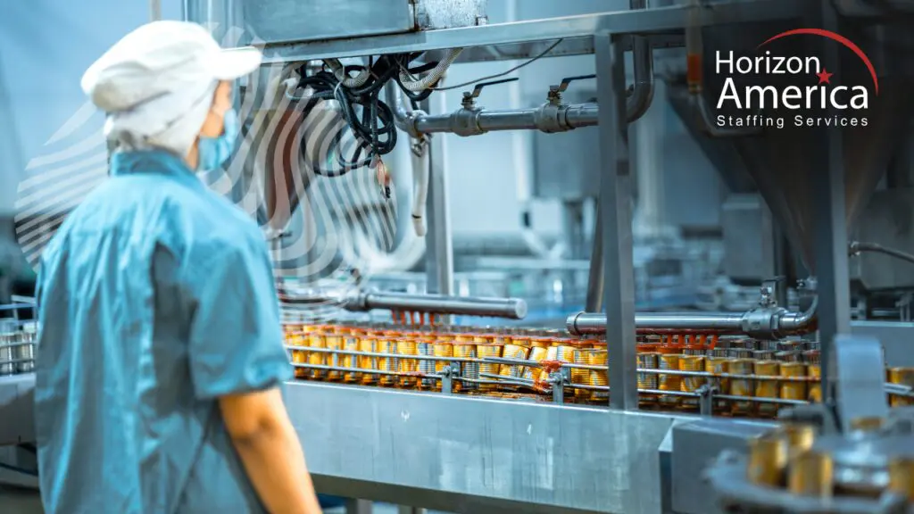 A food manufacturing worker in a hairnet, face mask, and blue uniform monitors canned goods moving along a production line in a food processing facility
