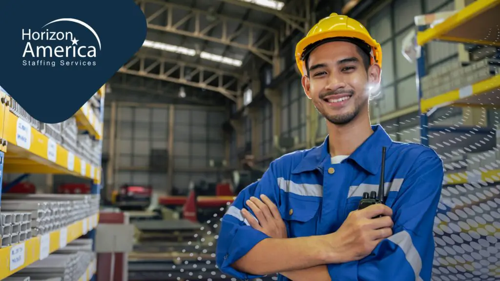 Smiling warehouse worker in a blue uniform and yellow hard hat standing with arms crossed in an industrial facility