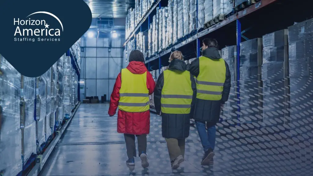 Three workers in high-visibility vests and winter jackets walking through a cold storage job environment lined with shelving and pallets.