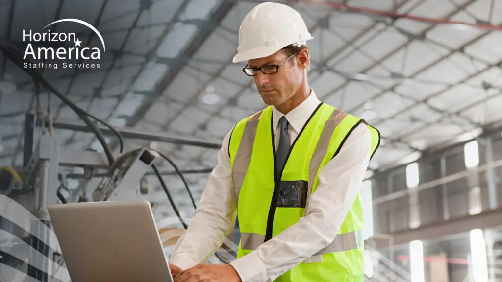 Warehouse manager in a high-visibility vest and hard hat reviewing data on a laptop while conducting a distribution staffing diagnostic inside a distribution facility.