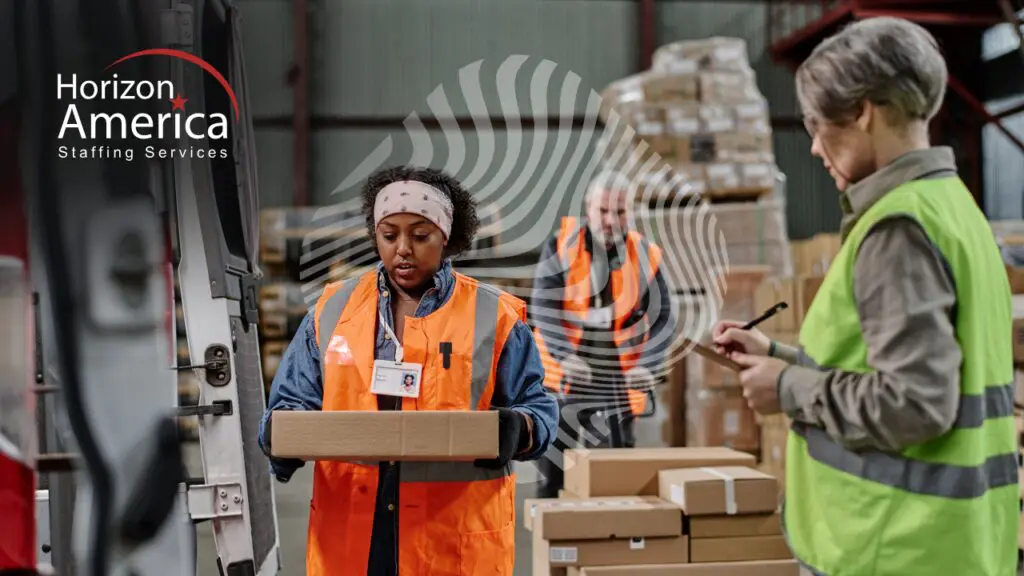 Distribution worker in a safety vest carrying a package while a supervisor reviews a clipboard in a warehouse.