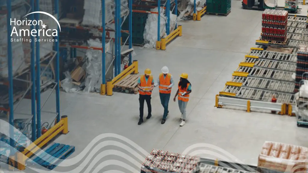 Three warehouse workers in safety vests and hard hats walk together in a warehouse