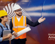 An executive wearing a safety vest and hard hat speaks to a woman in overalls and hard hat holding a tablet