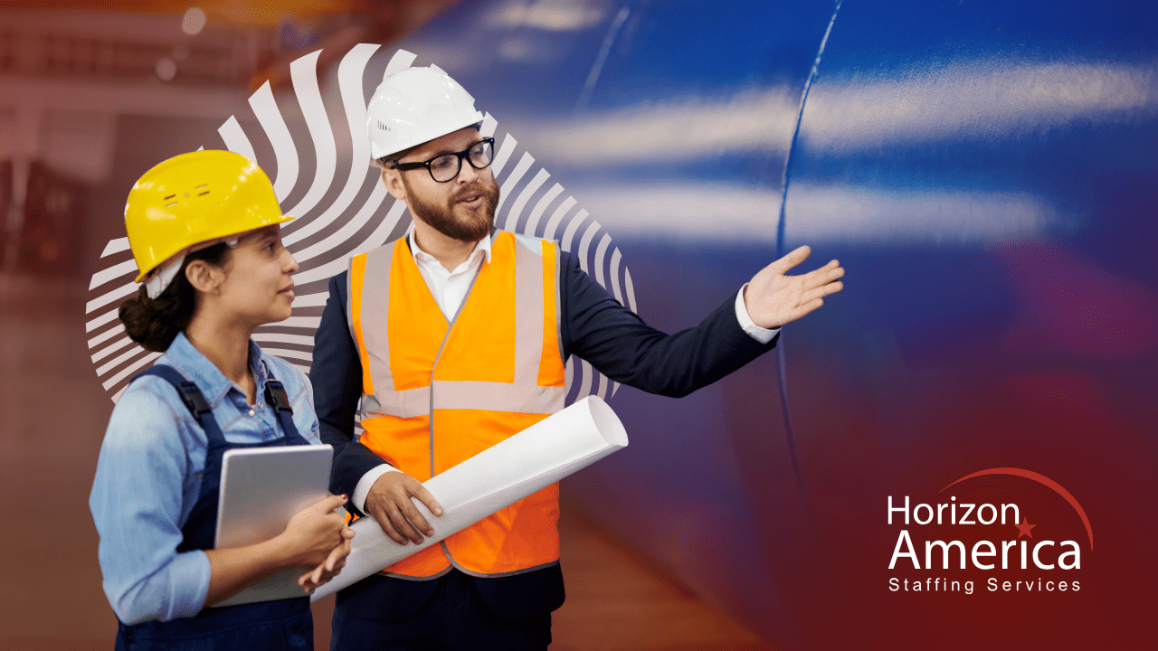 An executive wearing a safety vest and hard hat speaks to a woman in overalls and hard hat holding a tablet