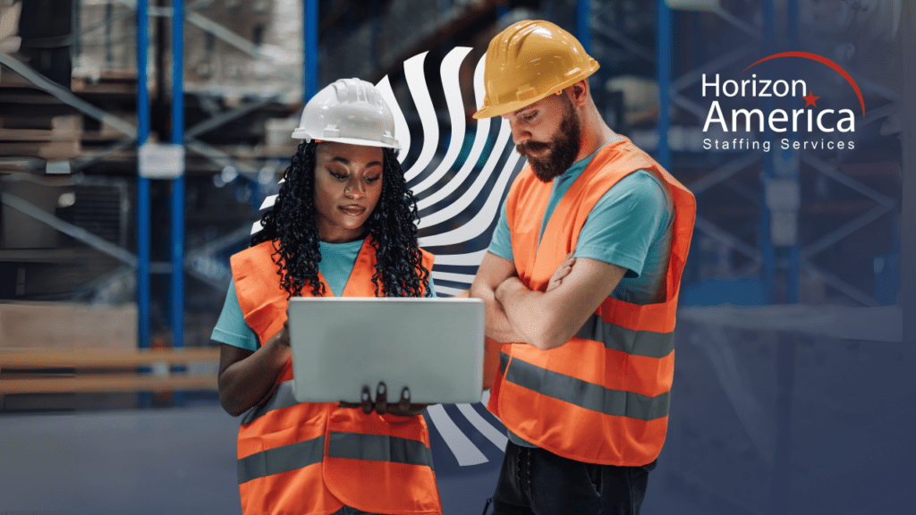 Two employees in safety vests and hard hats share a look at a laptop