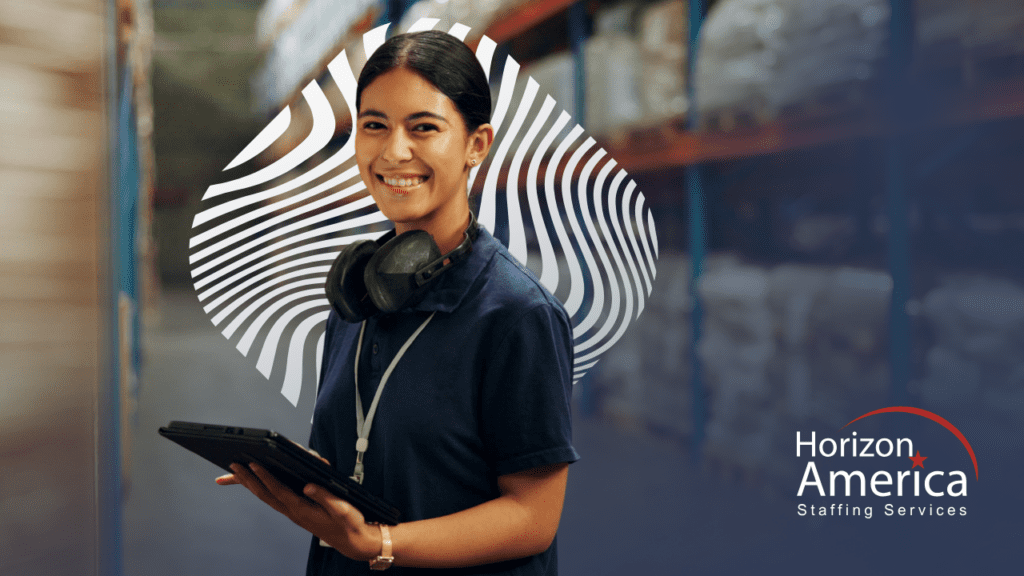 Light industrial employee stands smiling holding a tablet and wearing safety headphones